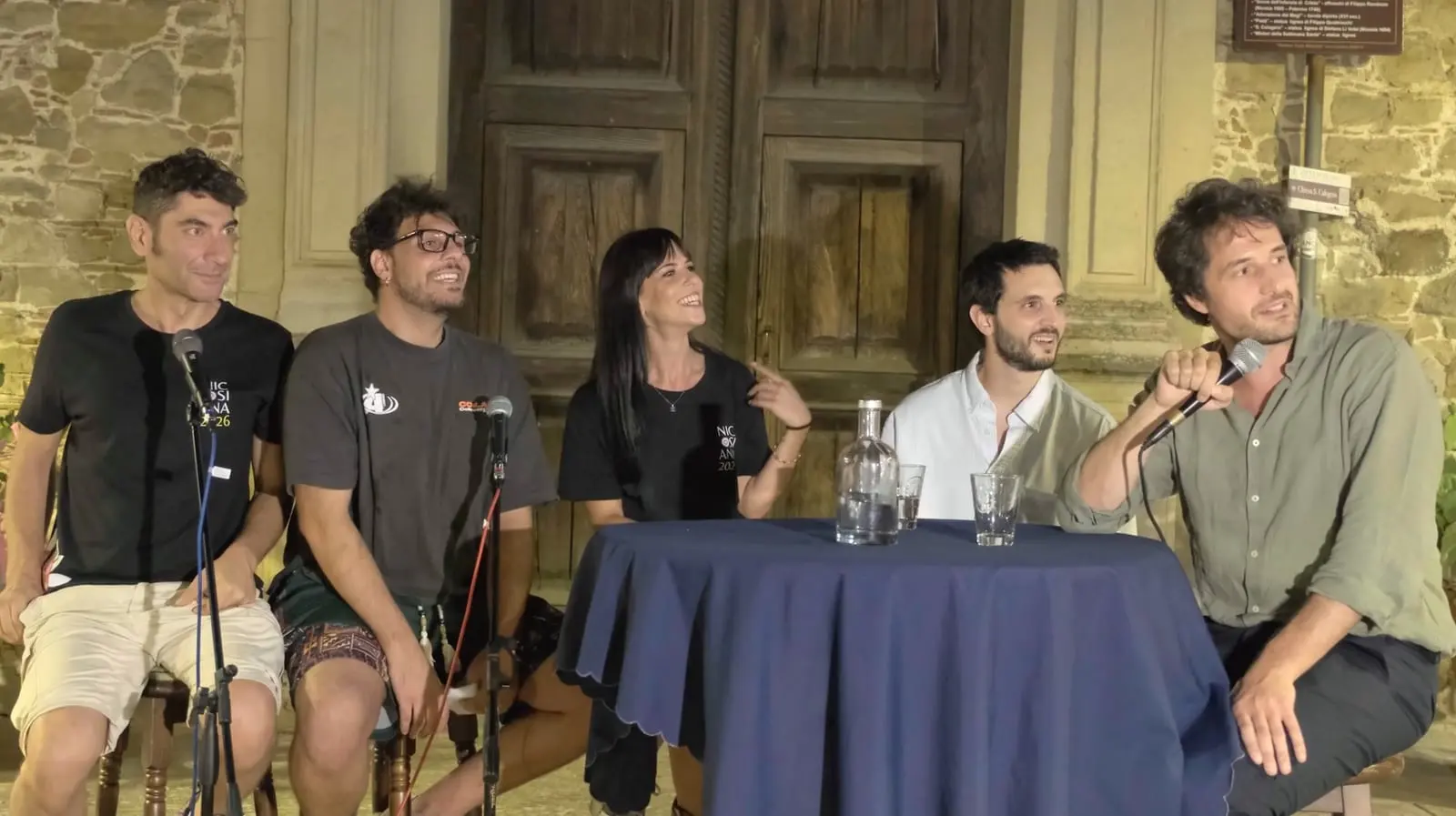 La registrazione della seconda puntata, davanti alla chiesa di San Calogero a Nicosia: cinque persone attorno al tavolino con la tovaglia blu, due con la maglietta nera di Nicosiana 2026; a destra un ospite parla al microfono.