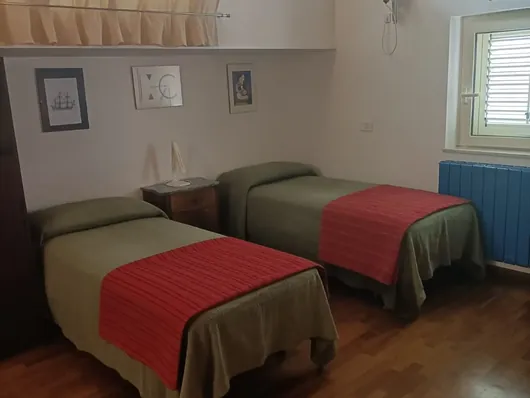 Due letti singoli con copriletti verdi e teli rossi, comodino e quadretti alla parete