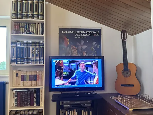 Angolo soggiorno con libreria bianca, televisore, chitarra classica e scacchiera su cassapanca
