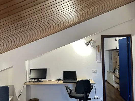 Angolo studio con scrivania a mensola, monitor, portatile e sedia sotto soffitto in legno